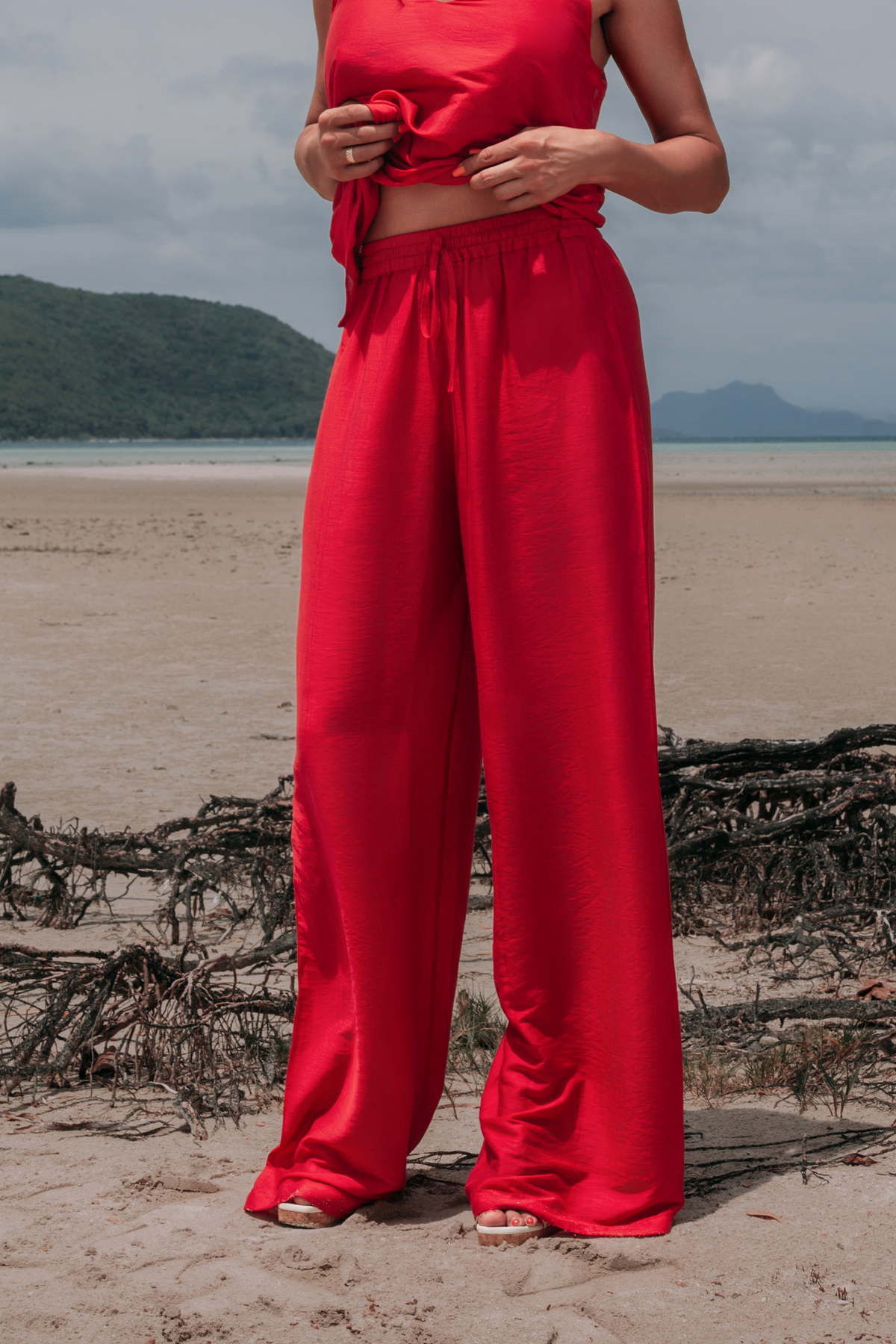 Carmine Fluid Pants by INNEVIA in deep red, front view of relaxed wide-leg trousers with a fluid drape on the beach