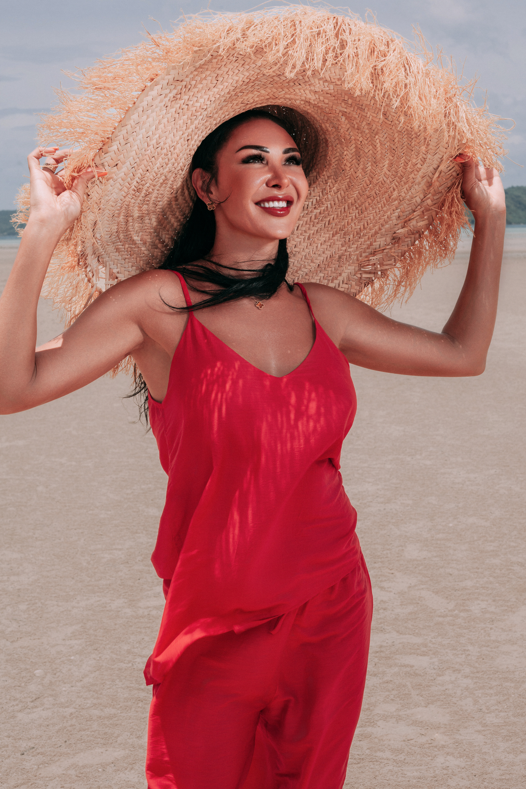 Carmine Top by INNEVIA in deep red with V-neckline and slim straps, styled with matching fluid trousers and an oversized straw hat on the beach