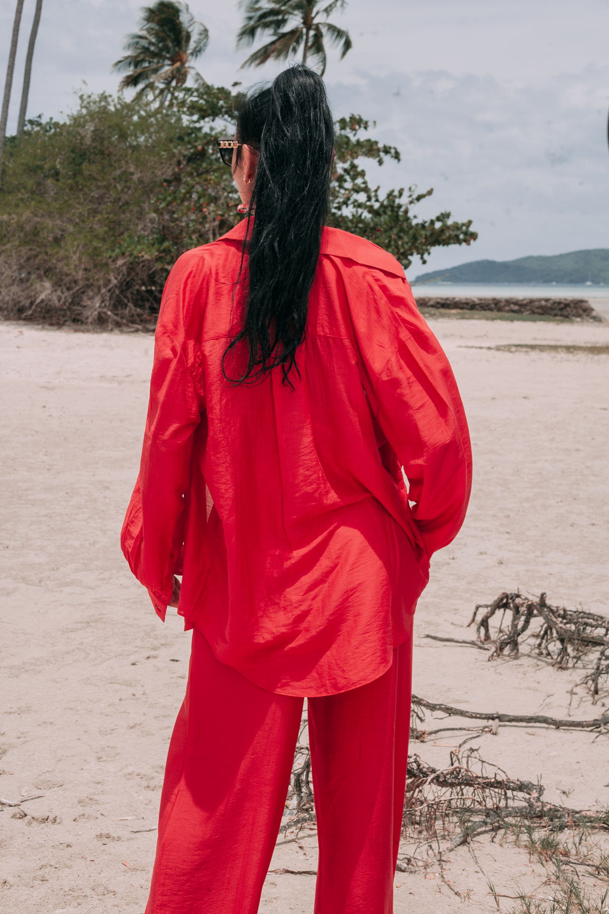 A woman standing on a sandy beach, viewed from behind, wearing a bright red matching two-piece outfit consisting of a long-sleeve collared shirt and wide-leg pants.