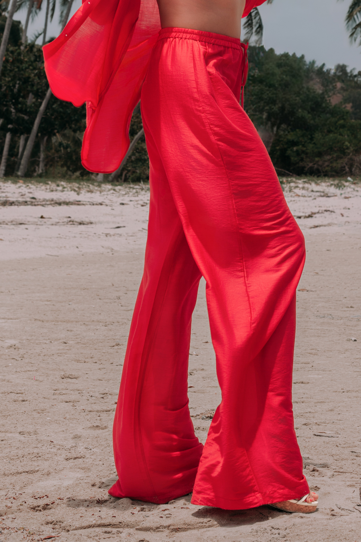 Carmine Fluid Pants by INNEVIA in motion, showing the flowing wide-leg silhouette and lightweight deep red fabric on the beach