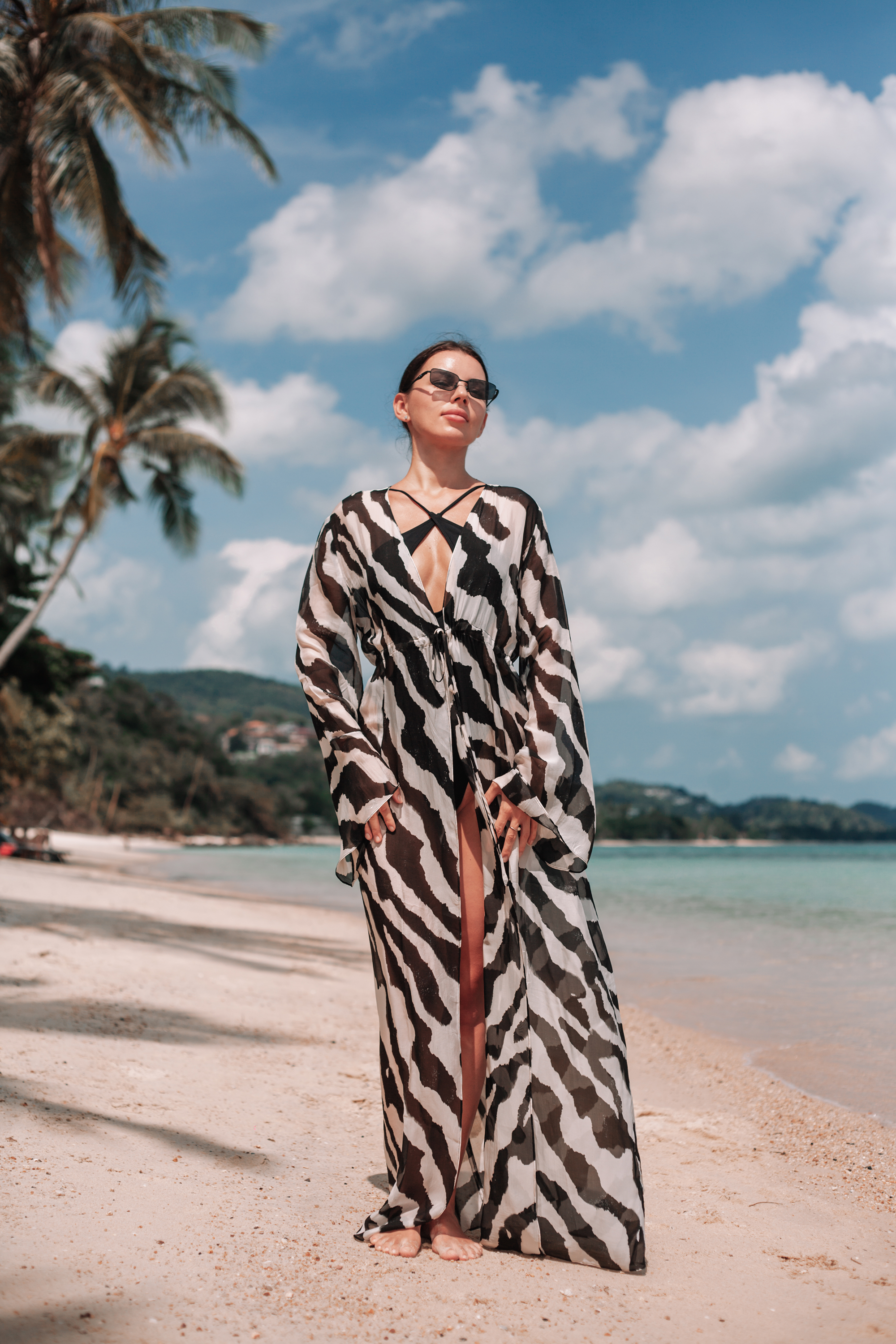 Woman in a zebra-striped dress standing on a beach with palm trees and blue sky in the background.