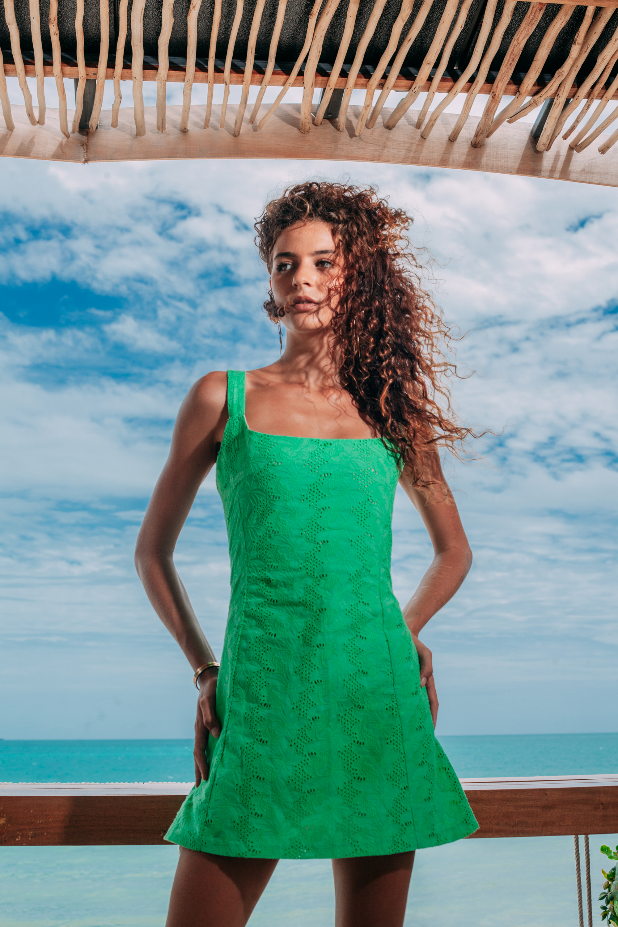 A woman with long curly brown hair models a bright green sleeveless mini dress with a textured, tonal floral pattern. She is standing on a wooden deck with a scenic ocean and cloudy sky background under a thatched roof.